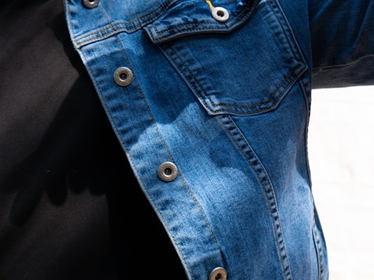 A close-up view of a person wearing a denim jacket with metal buttons and a black shirt underneath. The denim fabric shows various shades of blue, with visible stitching and a pocket on the chest. There are shadows cast on the jacket, indicating natural light.