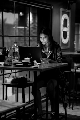 A woman is sitting alone in a dimly lit café, focused on her laptop. A glass with a stem and several other items, including a water bottle, are on the table. The setting is intimate and cozy, with large windows and minimal light.