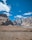Majestic mountains rise sharply against a clear blue sky, with some fluffy white clouds scattered above. The landscape is barren, with rocky textures and a sparse, earthy foreground. Large white letters on the ground spell out 'WELCOME TO PASSU', creating a striking contrast with the rugged terrain.