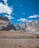 Majestic mountains rise sharply against a clear blue sky, with some fluffy white clouds scattered above. The landscape is barren, with rocky textures and a sparse, earthy foreground. Large white letters on the ground spell out 'WELCOME TO PASSU', creating a striking contrast with the rugged terrain.