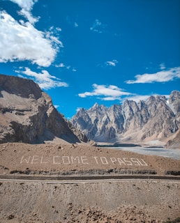 Majestic mountains rise sharply against a clear blue sky, with some fluffy white clouds scattered above. The landscape is barren, with rocky textures and a sparse, earthy foreground. Large white letters on the ground spell out 'WELCOME TO PASSU', creating a striking contrast with the rugged terrain.