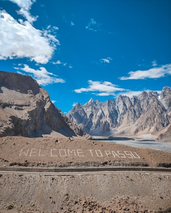 Majestic mountains rise sharply against a clear blue sky, with some fluffy white clouds scattered above. The landscape is barren, with rocky textures and a sparse, earthy foreground. Large white letters on the ground spell out 'WELCOME TO PASSU', creating a striking contrast with the rugged terrain.