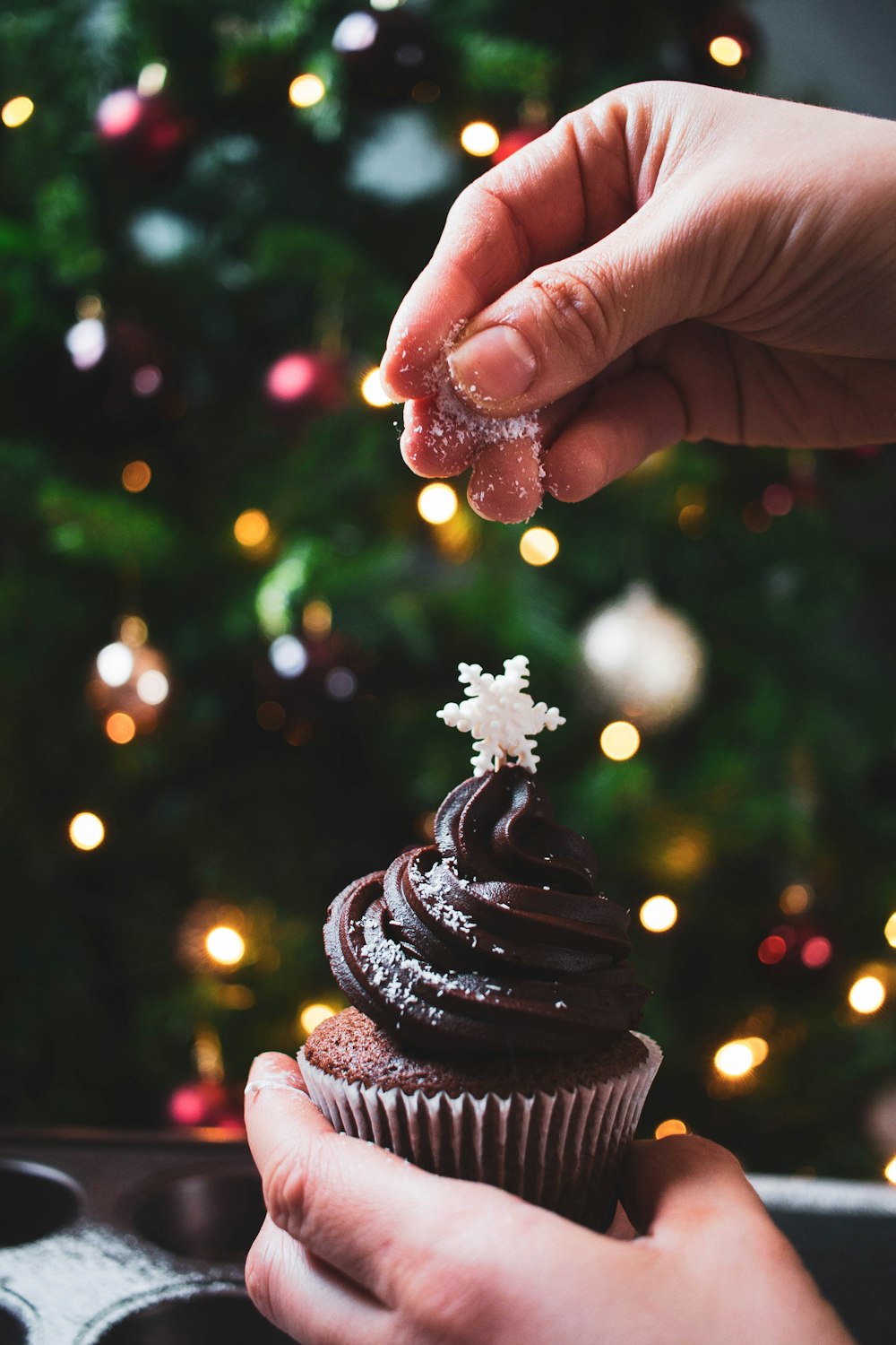 a person holding a chocolate cupcake in front of a christmas tree