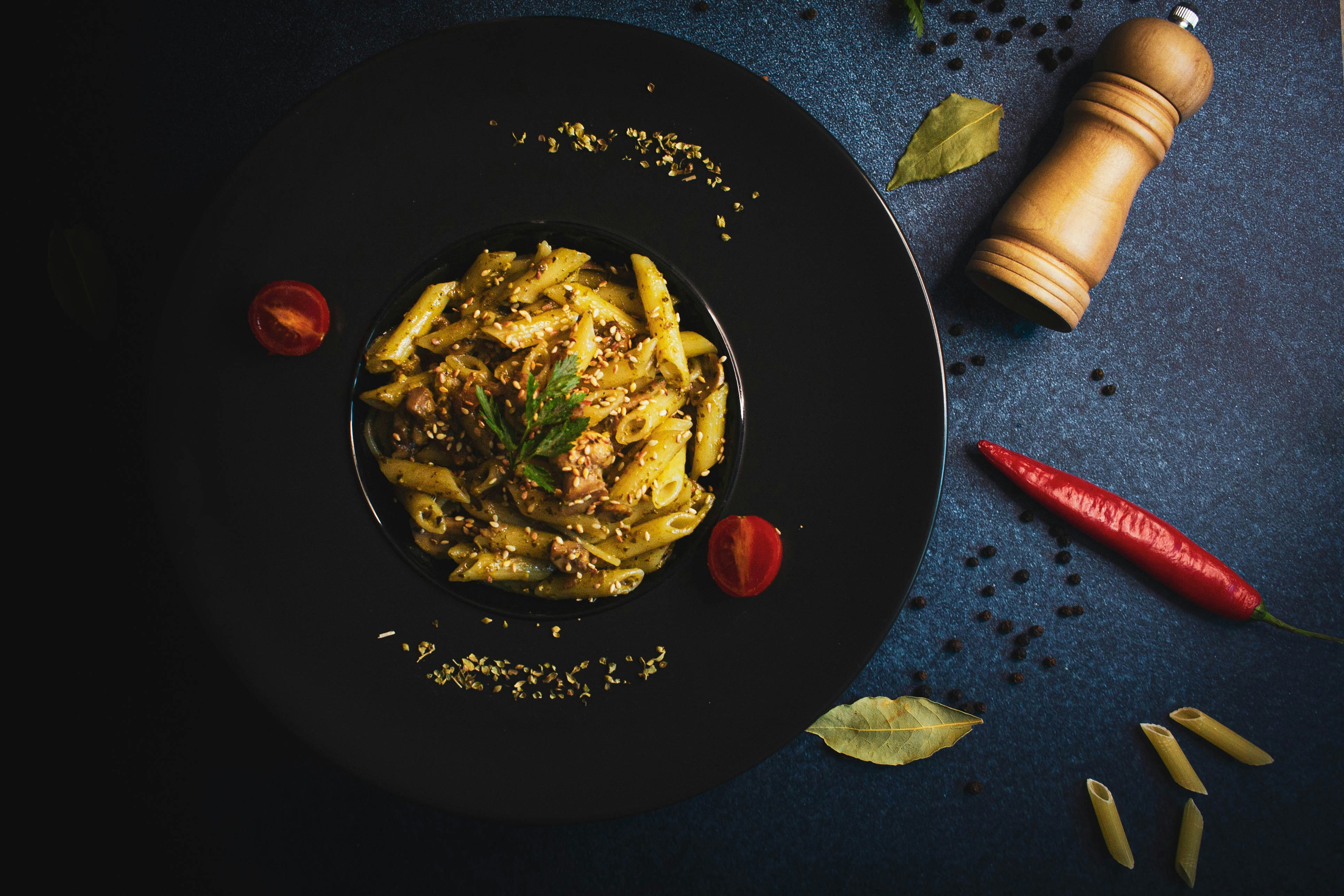 a black plate topped with pasta next to a pepper shaker, Served food on blue background