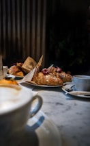 A cozy café setting featuring a marble table with a cappuccino in the foreground. Plates of pastries, including croissants topped with powdered sugar and berries, are arranged on the table. The background shows a warm, slightly blurred indoor ambiance with vertical wooden panels.