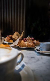 A cozy café setting featuring a marble table with a cappuccino in the foreground. Plates of pastries, including croissants topped with powdered sugar and berries, are arranged on the table. The background shows a warm, slightly blurred indoor ambiance with vertical wooden panels.