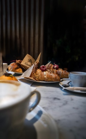 A cozy café setting featuring a marble table with a cappuccino in the foreground. Plates of pastries, including croissants topped with powdered sugar and berries, are arranged on the table. The background shows a warm, slightly blurred indoor ambiance with vertical wooden panels.