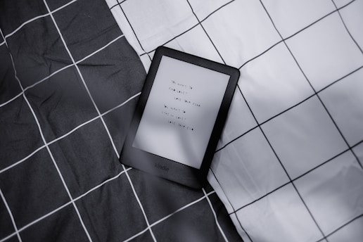 A Kindle device displaying text lies on a checkered fabric background with a monochrome color scheme. The text on the Kindle is partially visible.