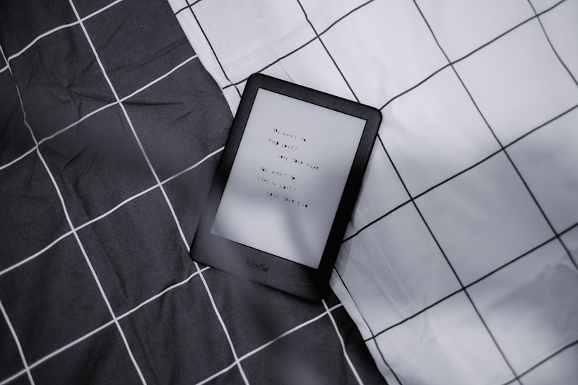 A Kindle device displaying text lies on a checkered fabric background with a monochrome color scheme. The text on the Kindle is partially visible.