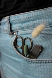 A pair of blue denim jeans with a pocket containing small scissors, a metal file, and a fuzzy cotton-like item. The jeans have a visible seam detail and stitching.