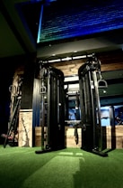 A gym setting featuring a large cable crossover machine with black metal frames against a wall of exposed bricks. The machine is situated on green artificial turf, and a wooden panel with a tree design is propped nearby. Ambient lighting casts blue and green hues onto the wall.