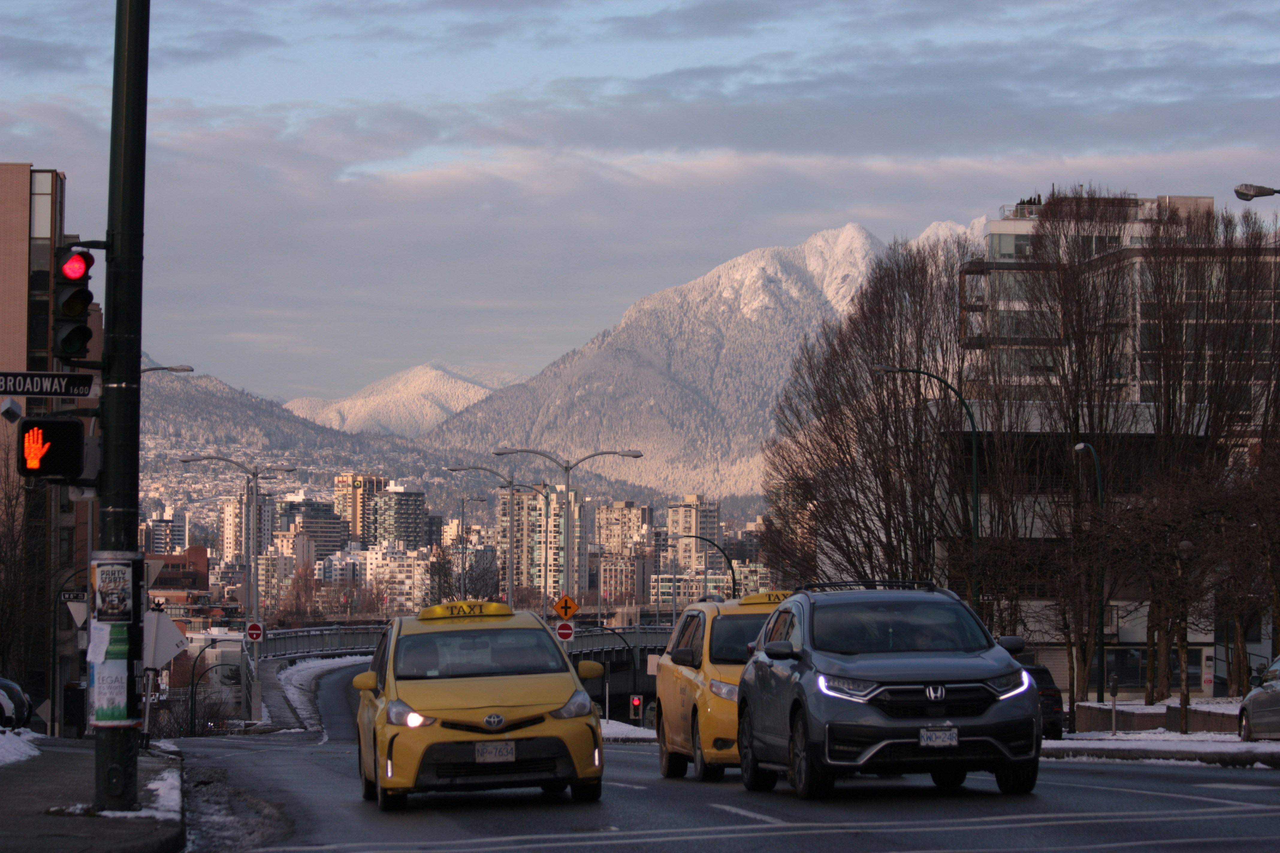 Yellow Vancouver taxis pull up to Broadway in winter with the snow-covered North Shore mountains behind them.