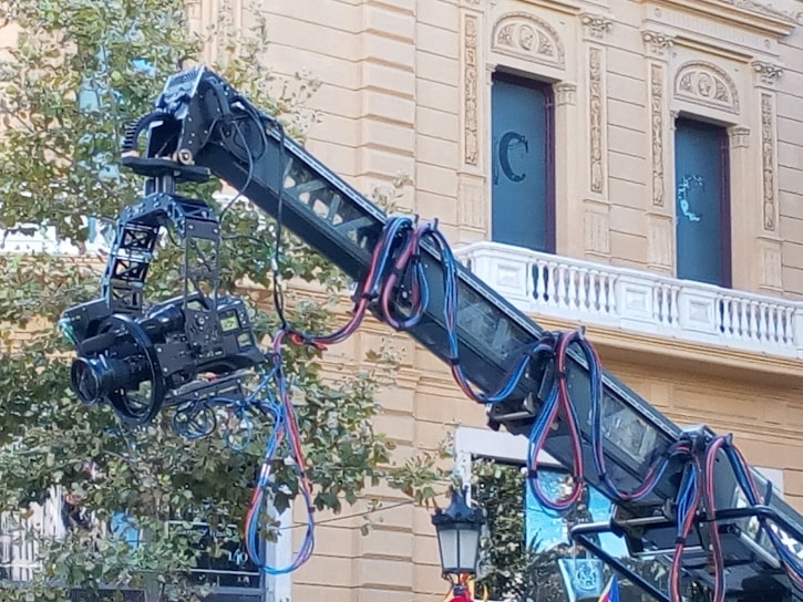 A large professional camera is mounted on a crane, framed against a classical building with ornate architectural details. Cables in various colors are connected to the camera and draped along the crane arm. Boxes and equipment can be seen in the background.