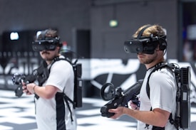 Two individuals are wearing virtual reality headsets and carrying gaming guns. Each person is equipped with a sturdy-looking backpack, possibly housing electronics needed for the VR experience. They are standing in an indoor environment, likely a virtual reality gaming arena, which has a modern and high-tech appearance. The background is slightly blurred, emphasizing the focus on the individuals.