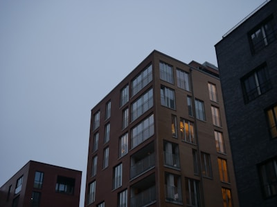 A group of modern apartment buildings with large windows against a backdrop of a clear, dimly lit sky. The central building is illuminated from within, suggesting occupancy. The structures are made of brick and concrete, giving a contemporary urban feel.
