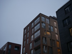 A group of modern apartment buildings with large windows against a backdrop of a clear, dimly lit sky. The central building is illuminated from within, suggesting occupancy. The structures are made of brick and concrete, giving a contemporary urban feel.