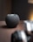 A small, dark gray spherical smart speaker with a mesh exterior sits on a wooden surface. The background features a blurred object in the foreground, possibly a camera or lens, with a warm light illuminating part of the scene.