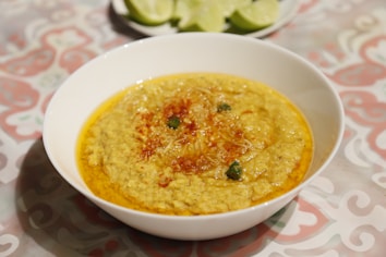 A bowl of yellow lentil soup is garnished with herbs and red spices, placed on a patterned tablecloth. In the background, lime slices rest on a plate.