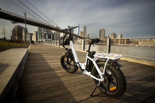 A white electric bike is parked on a wooden boardwalk with a scenic view of an urban skyline in the background. The scene is set during daylight, and the iconic structure of a large bridge spans across the upper part of the image. The foreground features a mix of metal railings and a paved path next to the wooden floor.