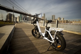 A white electric bike is parked on a wooden boardwalk with a scenic view of an urban skyline in the background. The scene is set during daylight, and the iconic structure of a large bridge spans across the upper part of the image. The foreground features a mix of metal railings and a paved path next to the wooden floor.