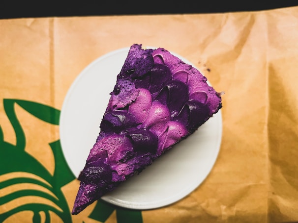 A slice of cake with vibrant purple frosting decorated in petal-like patterns. The cake sits on a white plate positioned on a paper surface with a green logo partially visible.