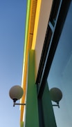 Bright and colorful building facade with a modern design and clean lines. The building features vibrant yellow and green accent colors, contrasting with a clear blue sky. A spherical lamp is mounted on the wall, adding an element of geometric interest.