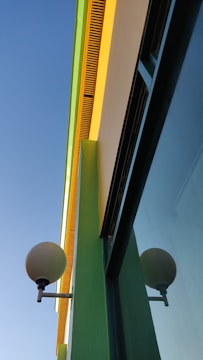 Bright and colorful building facade with a modern design and clean lines. The building features vibrant yellow and green accent colors, contrasting with a clear blue sky. A spherical lamp is mounted on the wall, adding an element of geometric interest.