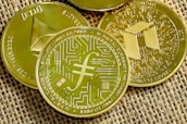 Several gold-colored cryptocurrency coins are displayed on a textured fabric surface. Each coin features distinct symbols and inscriptions related to digital currencies.