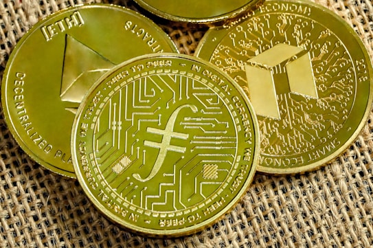 Several gold-colored cryptocurrency coins are displayed on a textured fabric surface. Each coin features distinct symbols and inscriptions related to digital currencies.