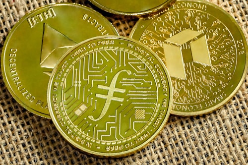 Several gold-colored cryptocurrency coins are displayed on a textured fabric surface. Each coin features distinct symbols and inscriptions related to digital currencies.