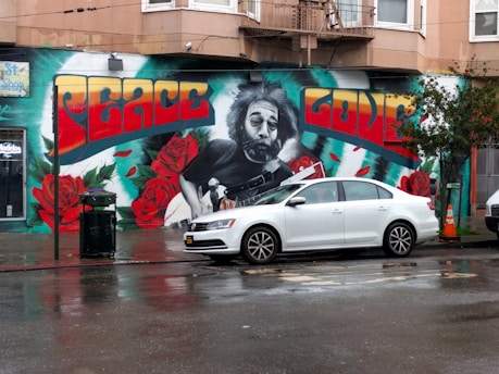 A vibrant mural on a city building features a large, detailed black-and-white portrait of a man playing a guitar, surrounded by vivid red roses and green leaves. The words 'Peace' and 'Love' are prominently displayed in large, colorful letters above the portrait. In front of the mural, a white car is parked on a wet street, reflecting the damp city environment.