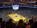 An indoor basketball arena filled with spectators. The court features a central logo and is surrounded by partially occupied tiered seating. A large scoreboard hangs above the center of the court, displaying the game score and time. Ambient lighting highlights the players on the court and the vibrant atmosphere of a live sports event.