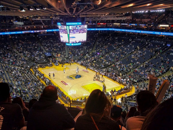 An indoor basketball arena filled with spectators. The court features a central logo and is surrounded by partially occupied tiered seating. A large scoreboard hangs above the center of the court, displaying the game score and time. Ambient lighting highlights the players on the court and the vibrant atmosphere of a live sports event.