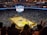 An indoor basketball arena filled with spectators. The court features a central logo and is surrounded by partially occupied tiered seating. A large scoreboard hangs above the center of the court, displaying the game score and time. Ambient lighting highlights the players on the court and the vibrant atmosphere of a live sports event.
