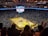 An indoor basketball arena filled with spectators. The court features a central logo and is surrounded by partially occupied tiered seating. A large scoreboard hangs above the center of the court, displaying the game score and time. Ambient lighting highlights the players on the court and the vibrant atmosphere of a live sports event.