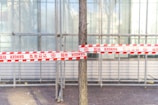 A tree is tied with red and white danger tape in front of a construction scaffolding or a glass structure. The tape has multiple warning signs and text in various languages. The ground is paved with bricks.