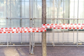 A tree is tied with red and white danger tape in front of a construction scaffolding or a glass structure. The tape has multiple warning signs and text in various languages. The ground is paved with bricks.