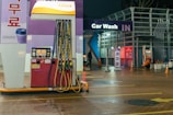 An outdoor car wash facility at night is illuminated by artificial lighting. In the foreground, there is a fuel dispenser station with multiple hoses and a payment terminal. The building has signage indicating 'Car Wash IN' with glowing letters. Cleaning equipment and supplies are visible on the right side. A person can be seen in the background setting up or handling cleaning materials.