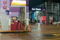 An outdoor car wash facility at night is illuminated by artificial lighting. In the foreground, there is a fuel dispenser station with multiple hoses and a payment terminal. The building has signage indicating 'Car Wash IN' with glowing letters. Cleaning equipment and supplies are visible on the right side. A person can be seen in the background setting up or handling cleaning materials.