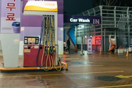An outdoor car wash facility at night is illuminated by artificial lighting. In the foreground, there is a fuel dispenser station with multiple hoses and a payment terminal. The building has signage indicating 'Car Wash IN' with glowing letters. Cleaning equipment and supplies are visible on the right side. A person can be seen in the background setting up or handling cleaning materials.