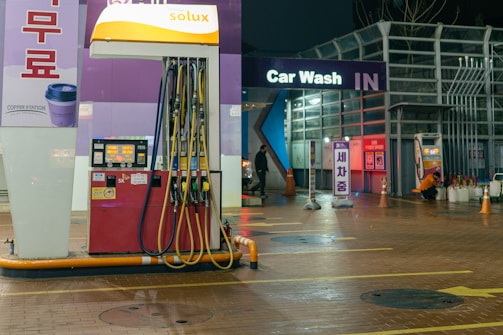 An outdoor car wash facility at night is illuminated by artificial lighting. In the foreground, there is a fuel dispenser station with multiple hoses and a payment terminal. The building has signage indicating 'Car Wash IN' with glowing letters. Cleaning equipment and supplies are visible on the right side. A person can be seen in the background setting up or handling cleaning materials.