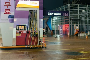 An outdoor car wash facility at night is illuminated by artificial lighting. In the foreground, there is a fuel dispenser station with multiple hoses and a payment terminal. The building has signage indicating 'Car Wash IN' with glowing letters. Cleaning equipment and supplies are visible on the right side. A person can be seen in the background setting up or handling cleaning materials.