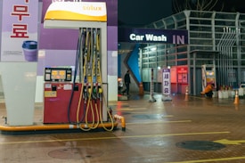 An outdoor car wash facility at night is illuminated by artificial lighting. In the foreground, there is a fuel dispenser station with multiple hoses and a payment terminal. The building has signage indicating 'Car Wash IN' with glowing letters. Cleaning equipment and supplies are visible on the right side. A person can be seen in the background setting up or handling cleaning materials.