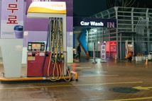 An outdoor car wash facility at night is illuminated by artificial lighting. In the foreground, there is a fuel dispenser station with multiple hoses and a payment terminal. The building has signage indicating 'Car Wash IN' with glowing letters. Cleaning equipment and supplies are visible on the right side. A person can be seen in the background setting up or handling cleaning materials.