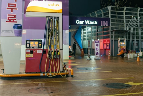 An outdoor car wash facility at night is illuminated by artificial lighting. In the foreground, there is a fuel dispenser station with multiple hoses and a payment terminal. The building has signage indicating 'Car Wash IN' with glowing letters. Cleaning equipment and supplies are visible on the right side. A person can be seen in the background setting up or handling cleaning materials.