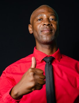 A person wearing a red shirt and black tie gives a thumbs up gesture. The background is dark, emphasizing the vivid color of the shirt and the expression of approval.
