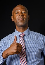 A man in a blue checkered shirt and a striped tie gives a thumbs up. The background is dark, and the lighting highlights his expression, which appears confident.