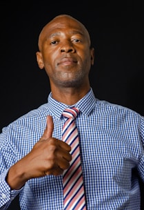 A man in a blue checkered shirt and a striped tie gives a thumbs up. The background is dark, and the lighting highlights his expression, which appears confident.