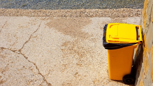 A yellow garbage bin with a black liner is positioned next to a wall on a paved surface. The pavement is cracked and weathered, and the bin casts a shadow on the ground. In the background, there is a section of rough, pebbled texture, possibly indicating a beach or riverside.