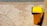 A yellow garbage bin with a black liner is positioned next to a wall on a paved surface. The pavement is cracked and weathered, and the bin casts a shadow on the ground. In the background, there is a section of rough, pebbled texture, possibly indicating a beach or riverside.